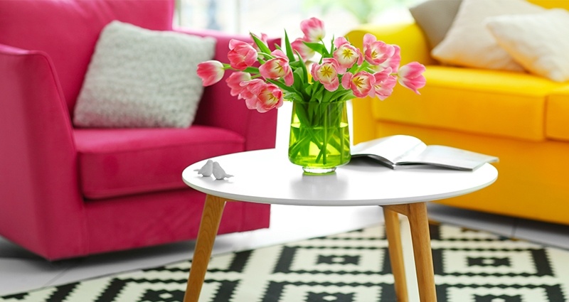 La photo d’une table avec une vase des tulipes roses sur un tapis noir et blanc, au fond un fauteuil amarante et un canapé jaune.
